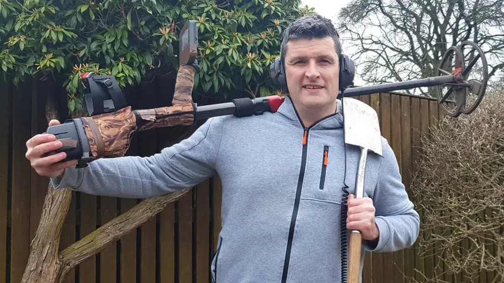 A male detectorist holding a metal-detector and a shovel. Pictures appears to be taken in a backyard.