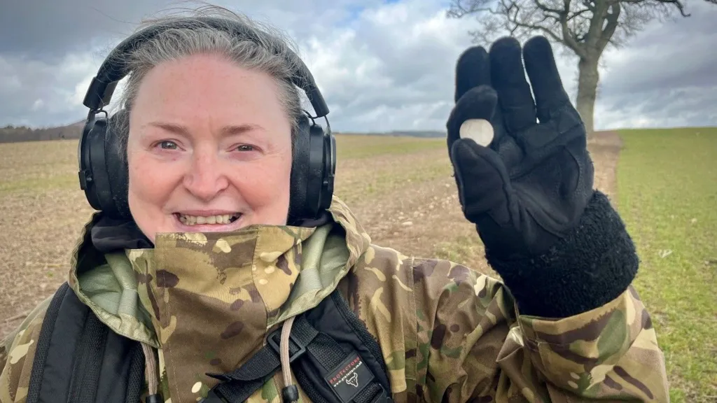 A female detectorist holding up a coin. The picture is taking in a field.