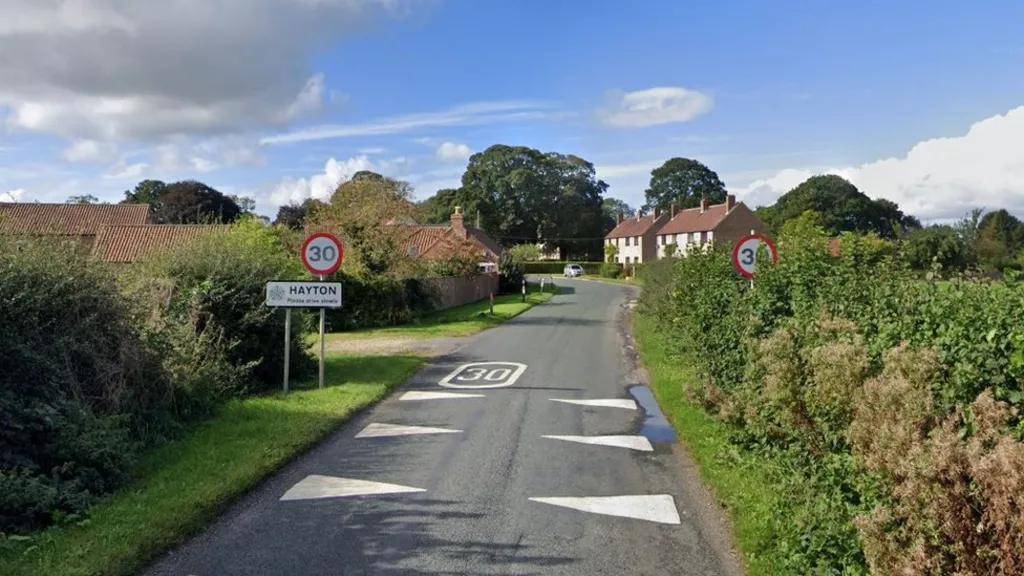 View of a country road with a village in the background. Road sign reads Hayton