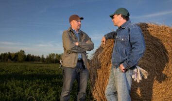 Two men talking while standing beside a haystack.